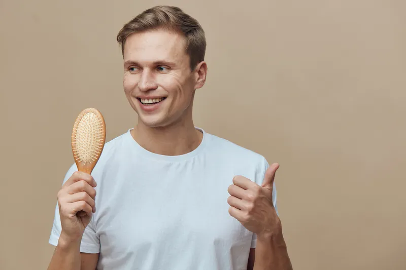 Smiling man with brown hair wearing light blue t-shirt holding hairbrush and showing thumbs up gesture, isolated on beige background, casual lifestyle concept.