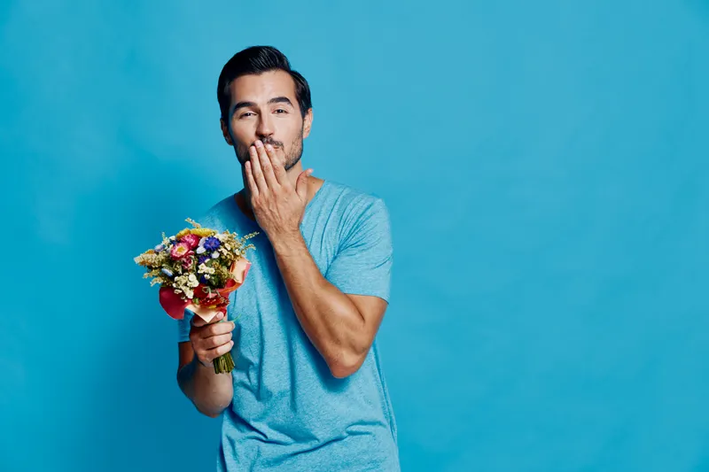 Handsome smiling man holding a bouquet of flowers with a playful expression, isolated on a bright blue background. Romantic gesture, happiness concept.