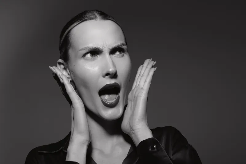 A shocked Caucasian woman with fair skin holds her face with both hands, mouth open in surprise or disbelief. Black and white studio portrait with dramatic lighting and expression.