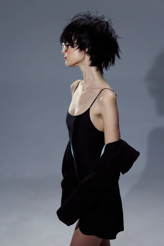 Young woman with short black hair wearing a black dress and glasses, standing against a neutral gray background, stylish and modern look.