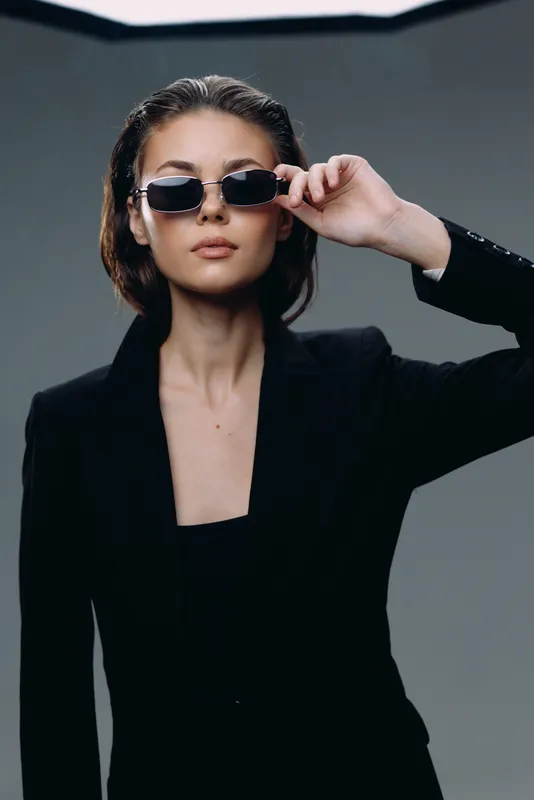 Stylish woman in black suit wearing sunglasses with a confident expression and modern hairstyle in a dark studio setting