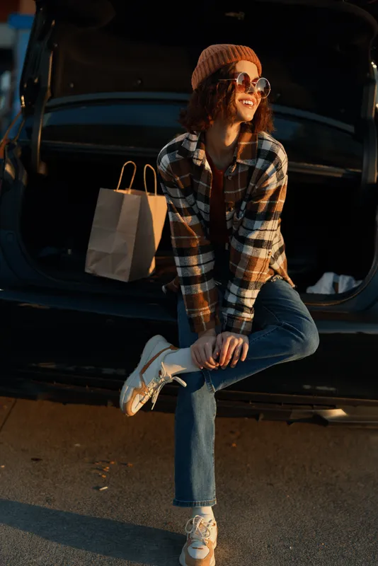 Woman car shopping smile sunglasses beanie plaid trunk candid lifestyle portrait of a joyful shopper sitting on an open car trunk at golden hour glow, authenticity and emotional storytelling.