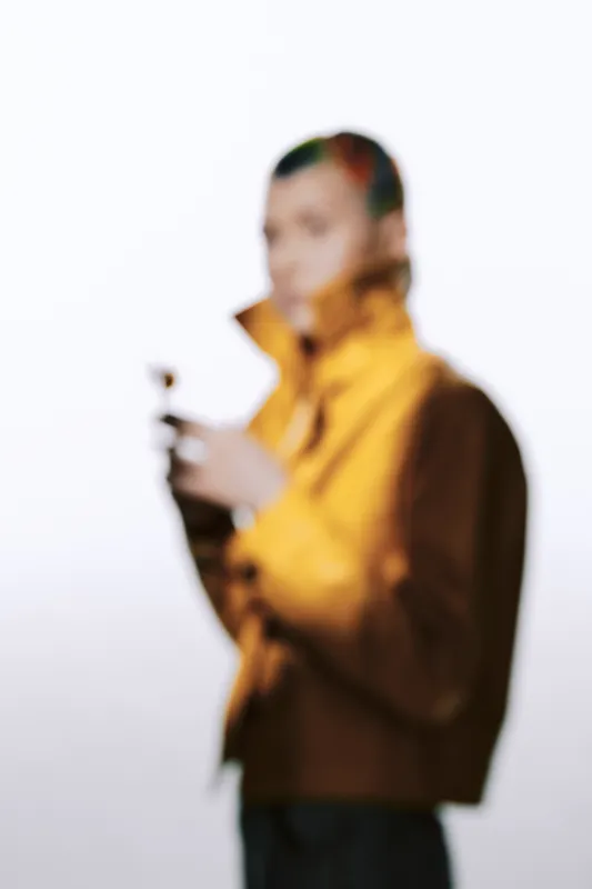 Blurred woman wearing yellow jacket holding an object, minimalistic background, soft lighting, contemporary casual style, artistic composition.