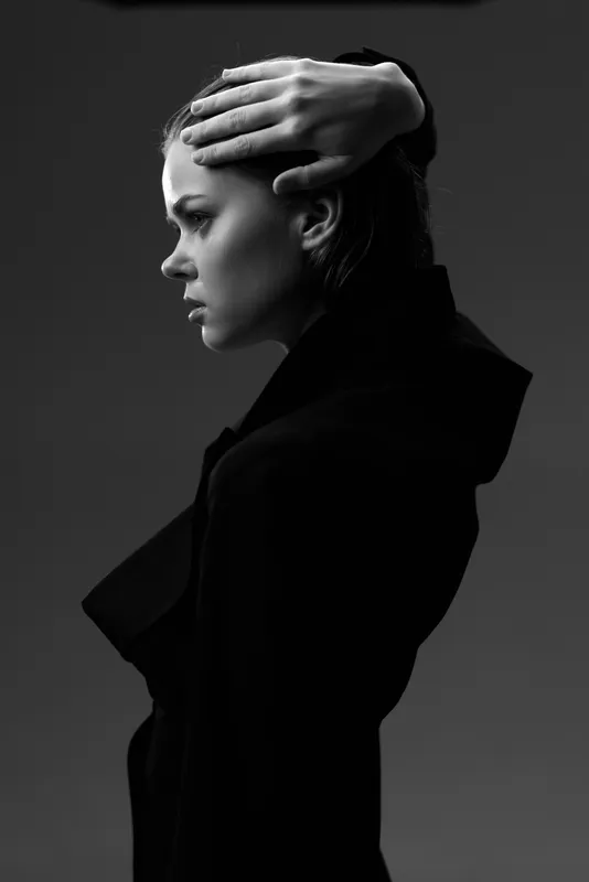 Black and white portrait of a young woman with contemplative expression in dark clothing against plain background.