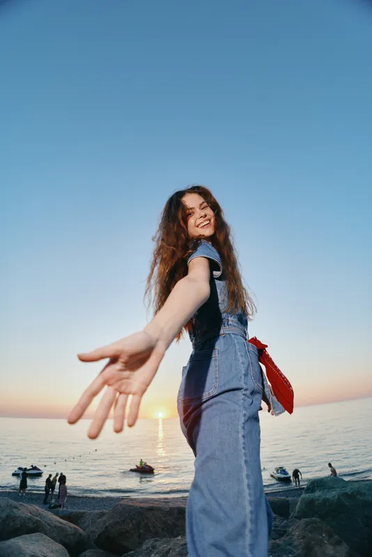lifestyle woman in modern boho-western denim street style reaches out happily at sunset by the ocean with warm film color tones and relaxed summer vibe