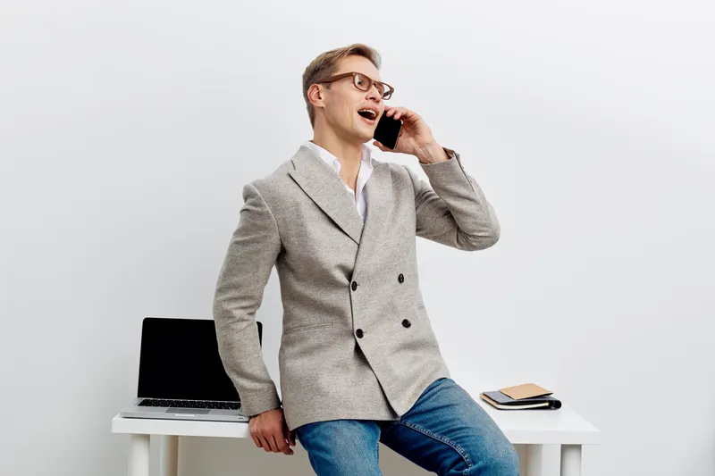 Smiling woman wearing gray blazer and glasses talking on mobile phone while leaning on white desk with laptop and notepad isolated on plain gray background, studio portrait. People lifestyle concept