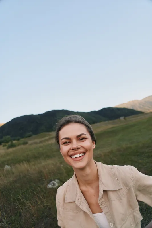 Smiling woman with natural makeup stands outdoors in casual shirt on green grassy hill under clear blue sky enjoying fresh air and peaceful nature landscape.