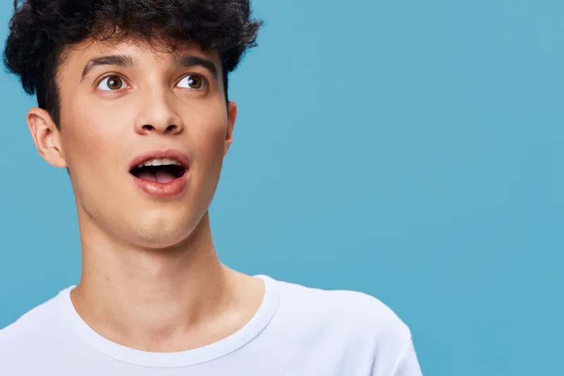surprised man with curly hair wearing white shirt looking amazed on blue background, youthful expression, casual style, studio portrait lighting