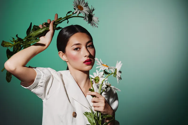 Elegant woman in white outfit holds fresh flowers with green background. Natural makeup and red lips create a stylish beauty fashion concept with soft natural light.