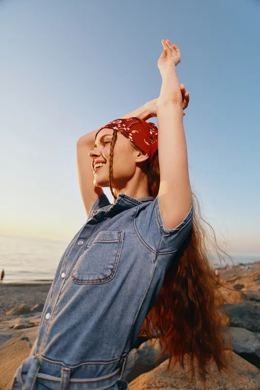 Lifestyle woman wearing modern boho-western denim street style with a red bandana, enjoying sunset by the rocky seaside, film color tones enhancing natural mood.
