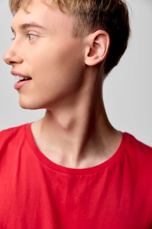 Profile view of a young man in a red shirt, captured with soft studio lighting, showcasing a relaxed smile and clean lines for fashion, lifestyle, and character stock imagery.