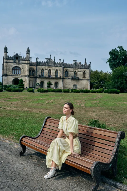 Woman sits on a bench in a park with a castle in the background, enjoying outdoor leisure under sunny skies, a serene moment of travel and exploration