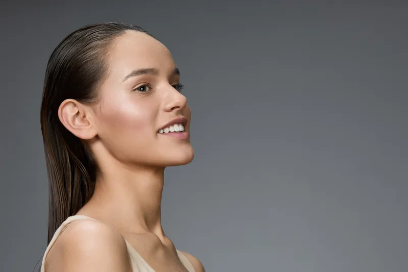 Beautiful young woman with long hair smiling against a grey background, showcasing healthy skin and elegance, perfect for beauty and skincare concepts