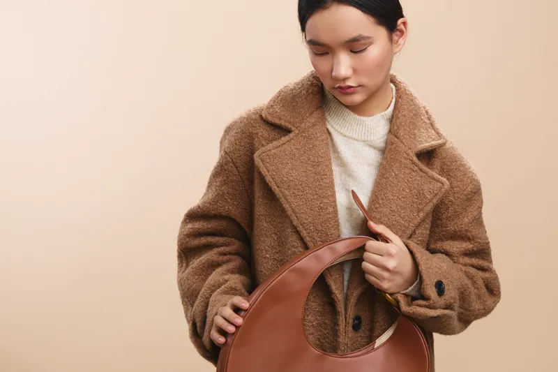 Asian woman in a chic brown coat examines a large leather bag against a neutral backdrop, showcasing casual elegance and contemporary fashion style in a poised studio setting.