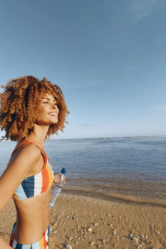 Smiling woman with curly hair wearing striped swimsuit, standing on a sandy beach by the ocean, gentle ocean waves nearby, enjoying summer vacation, outdoors, bright natural light, clear sky