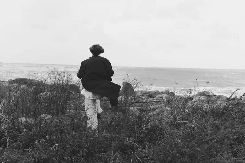 Man walking along a rugged coastal landscape in black and white, capturing solitude by the ocean, windswept grasses and a vast sky over the shore.