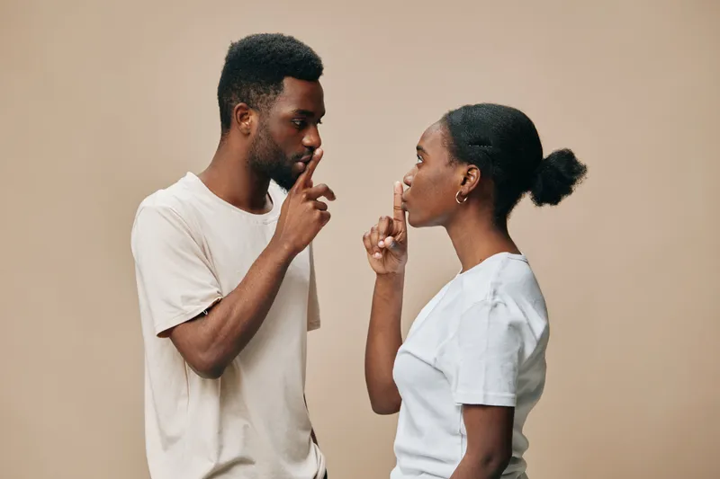 Two young adults silently communicating with finger gestures against a soft beige background, showcasing emotions of secrecy and understanding