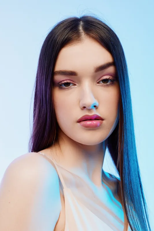 Young woman with long straight hair and glossy lips poses against a light blue background. Her makeup highlights natural beauty and fashion elegance in a studio setting.