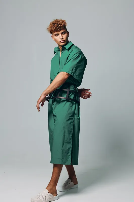 Young man with curly hair wearing green oversized shirt and matching wide shorts posing in studio with white slip-on shoes and black belt accessory on gray background.