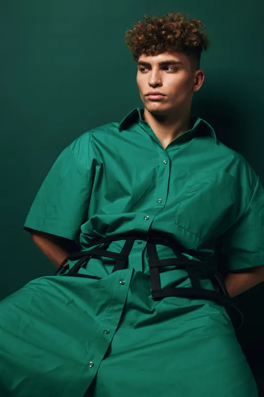 Portrait of a young man sitting and looking to the side wearing green pajamas with curly hair in a studio environment with green background. Male model with patterned haircut.