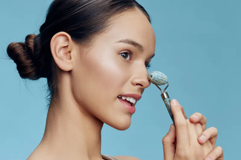 Young woman with smooth skin applying a facial roller for skincare, showcasing a radiant expression against a soft blue background