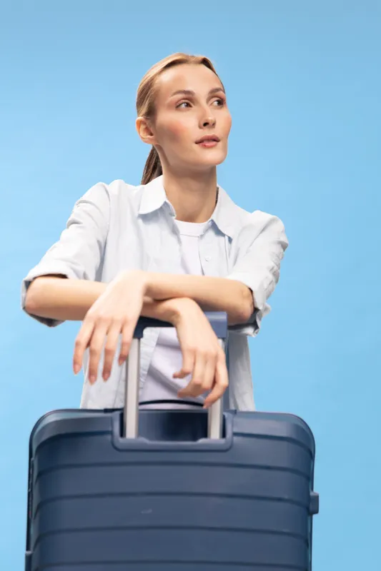 Young woman with suitcase leans on handle and looks away thoughtfully on blue background. Concept of travel, vacation, and planning a trip with calm mood.