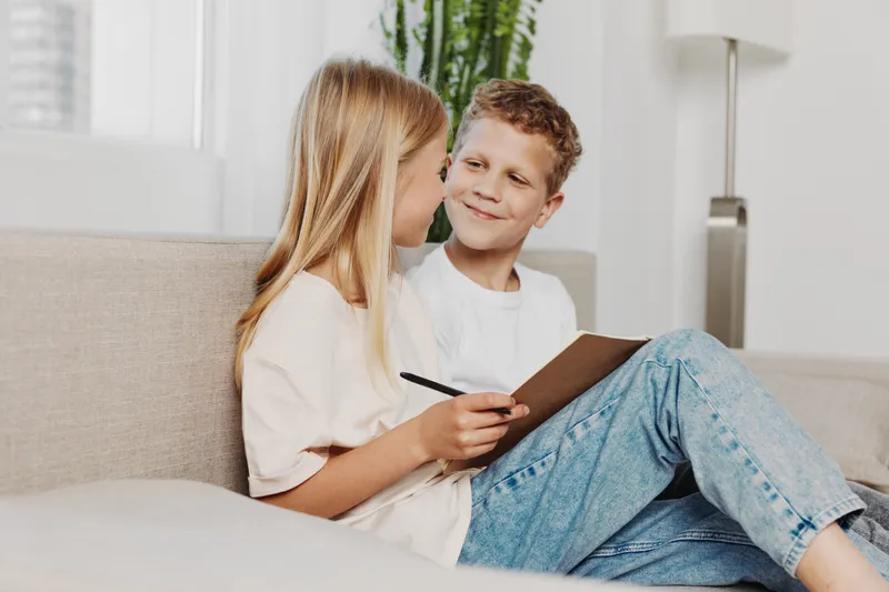 Two children sitting on sofa in bright living room, girl holding notebook and pen while boy looks at her smiling. Concept of childhood friendship, learning, and casual home environment.