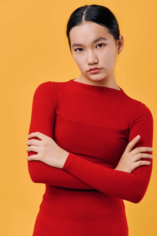 Young woman with dark hair wearing red long sleeve shirt standing with crossed arms on orange background. Portrait of serious confident female model with natural makeup and casual style.