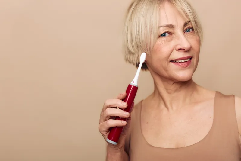 Smiling mature woman with short blonde hair holding red electric toothbrush near face on beige background. Concept of dental care, oral hygiene, wellness, and daily routine for seniors.
