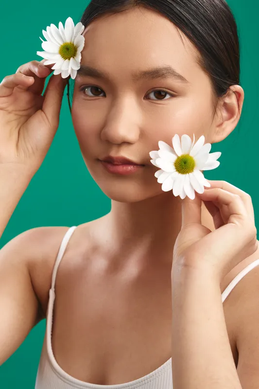 asian woman holds daisies near her face with a vibrant green background, a soft smile and calm expression that conveys freshness, natural beauty, and a gentle, dreamy mood.