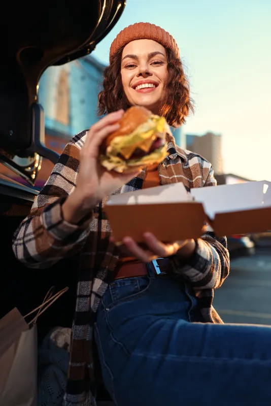 Woman holding burger and takeaway by car trunk, smile in urban evening light. Candid lifestyle portrait with authenticity, golden hour glow, mindful living and emotional storytelling.