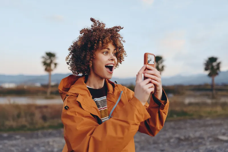 Woman lifestyle selfie outdoors smiling curly phone jacket travel recreation, laughing woman holds smartphone for selfie, orange jacket, coastal background, palm trees, bright sky
