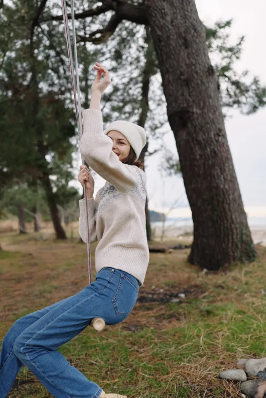 Woman on an outdoor swing in a park, wearing a cozy knit sweater and beanie, smiling as she leans back among trees and green grass.