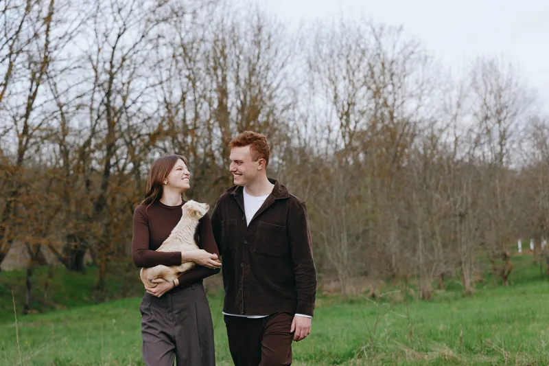 Casual couple outdoors with a goat kid, smiling and holding hands, in a green meadow under soft daylight, a wholesome lifestyle moment that feels natural, warm, intimate, and freely candid.