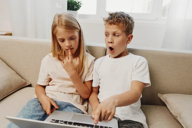 Curious boy and thoughtful girl sitting on sofa using laptop together at home. Children explore digital content with focused expressions in bright living room. Concept for learning and technology.