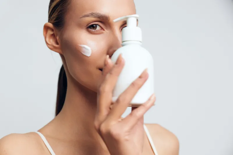 Young woman applying moisturizer cream on face holding white plastic bottle close to skin, skincare routine, natural beauty, clean fresh look, isolated background