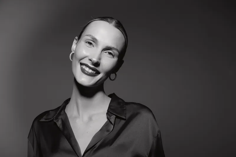 Smiling Caucasian woman with light skin and slicked back hair wears black silk shirt and hoop earrings, posing in studio with soft lighting for fashion portrait.