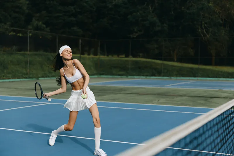 Energetic young woman playing tennis outdoors in sporty attire, exhibiting joy and enthusiasm on a sunny day, surrounded by green trees and blue tennis courts