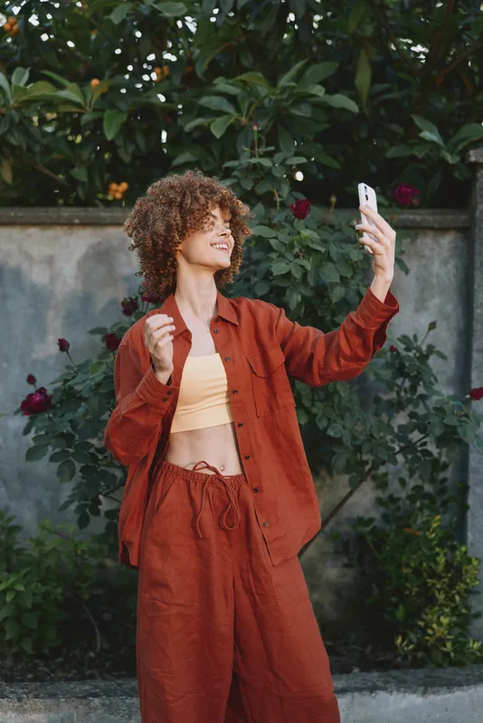 Smiling woman with curly hair takes a selfie outdoors, wearing rust colored casual outfit, garden background. Lifestyle moment, self expression, and cheerful energy. Natural light highlights texture