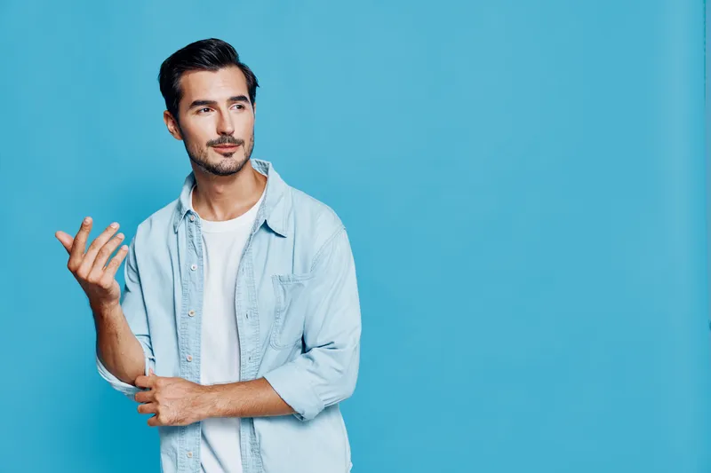 Stylish young man in casual outfit gestures thoughtfully while standing against a vibrant blue background, showcasing a modern lifestyle and personal expression.