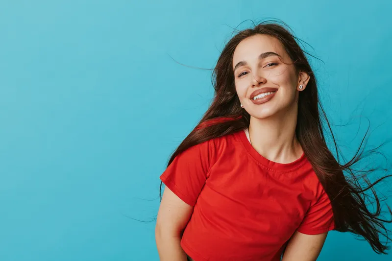 Young woman smiling and posing in red t shirt with long hair flowing on blue background. Portrait of happy female with natural makeup and earrings in casual style studio shot.