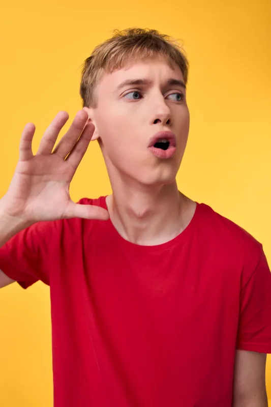 Shouting man in a red shirt gestures with his hand beside his face, delivering a loud, urgent message with expressive energy against a bright yellow background for marketing use