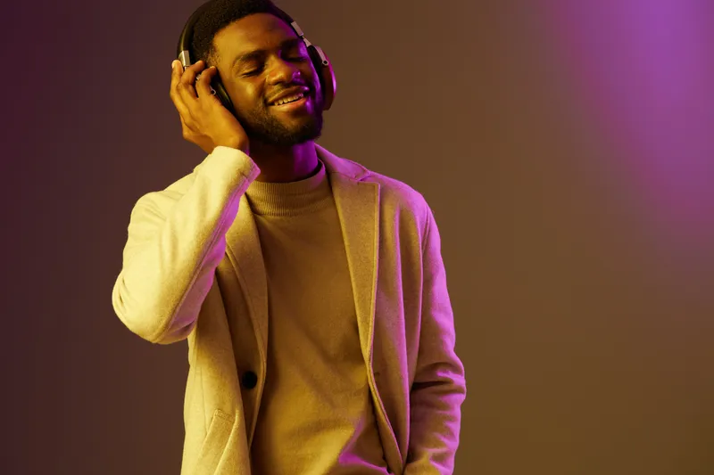 Happy Black man enjoying music with headphones, wearing a stylish outfit against a colorful backdrop, evoking a sense of joy and relaxation in a modern setting