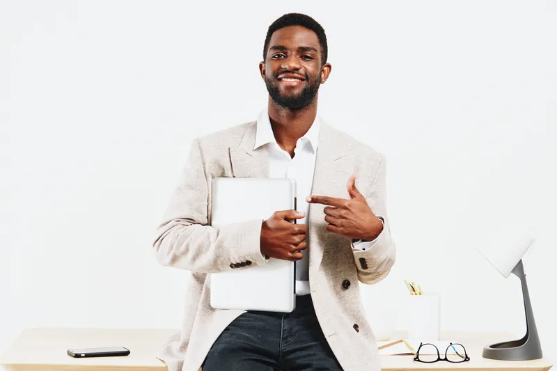 Confident smiling man in beige blazer holding laptop and pointing at it, looking at camera in modern office environment. People, professional, success, business, technology, workspace, professional at