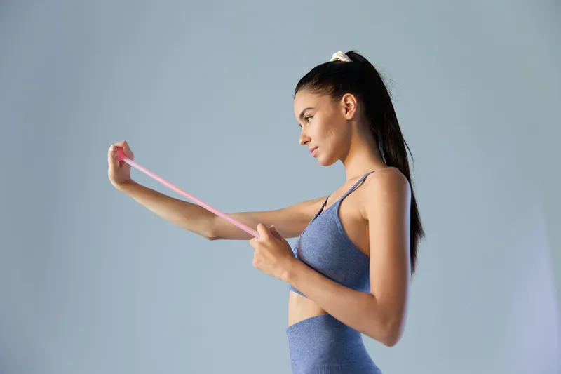 fit woman exercising with resistance band in blue sportswear, focused young female athlete training, indoor fitness, health and workout concept