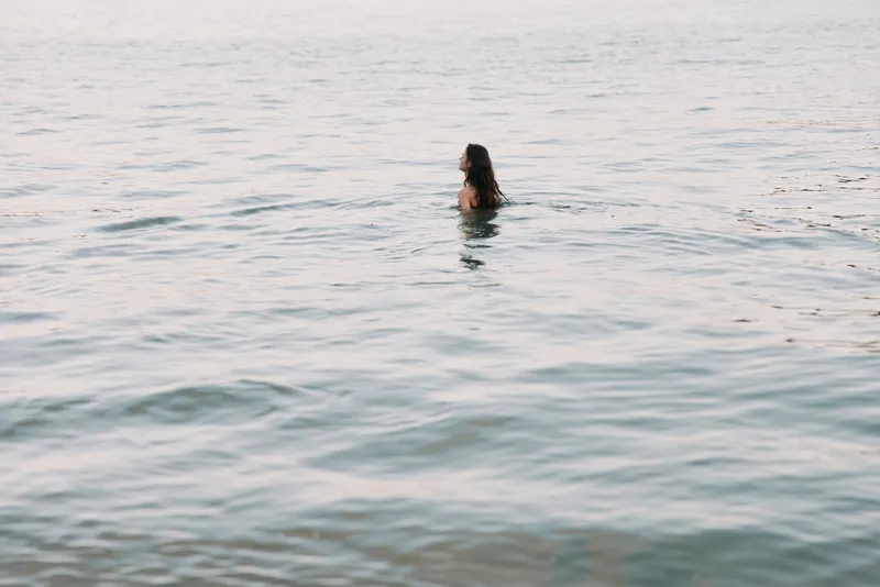 A solitary swimmer emerges from calm, glassy water, with the body partially visible as gentle ripples surround, creating a serene, contemplative aquatic moment.