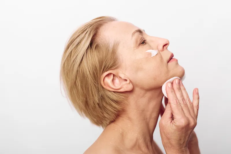 Mature woman with short blonde hair applying white facial cream with cotton pad on clean skin. Skincare routine concept focused on anti aging, beauty care, and natural skin health in studio.