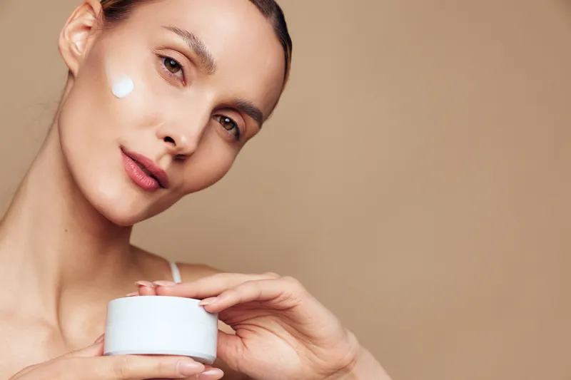 A young woman with clear skin applies white cream on her cheek while holding a white cosmetic jar against a beige background. Concept for skincare, beauty, and facial care.