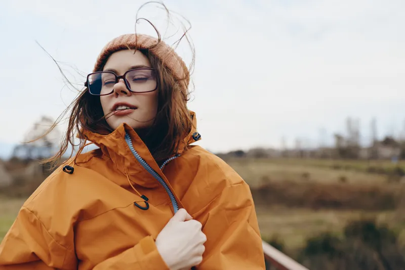 Woman lifestyle outdoors in an orange jacket and glasses, wind moves her hair, she adjusts the coat, countryside field background, soft daylight, casual travel portrait