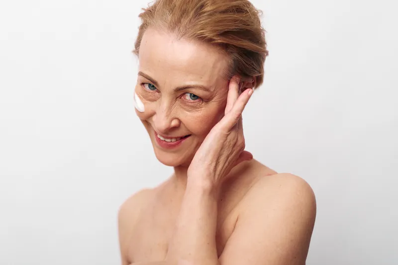 Smiling middle aged woman with bare shoulders applies white face cream on cheek. Studio shot on white background. Concept of skincare, anti aging, self care, and natural beauty.